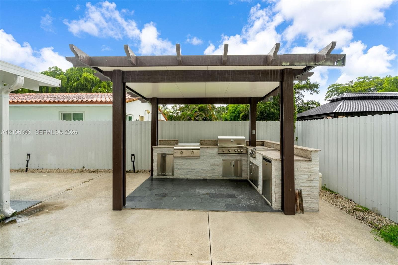 Covered outdoor kitchen featuring a pergola, stone-clad island, built-in stainless steel grill, beverage cooler, and slate-finish flooring