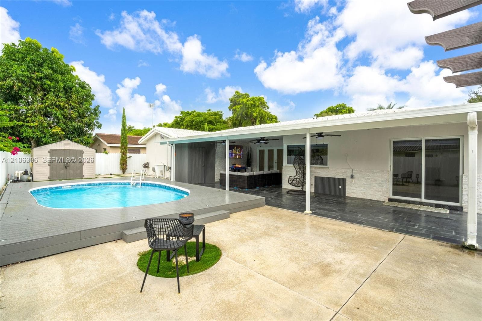 Backyard space featuring an above-ground swimming pool with a raised wood-finish deck, a concrete patio, a covered lanai with stone-style siding, and a detached storage structure