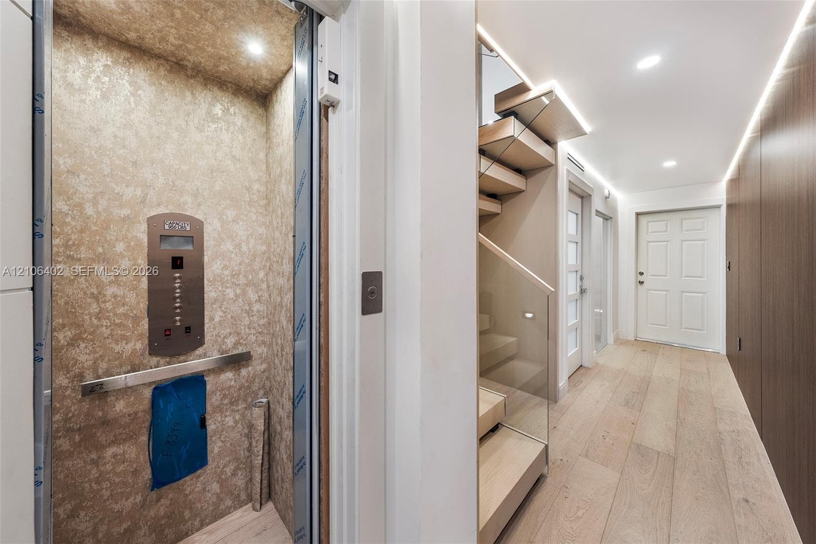 Private elevator featuring metallic-finish wall paneling and a stainless steel control panel, wood-finish flooring, staircase with glass railings and wood treads, recessed lighting, and wall-mounted linear lighting
