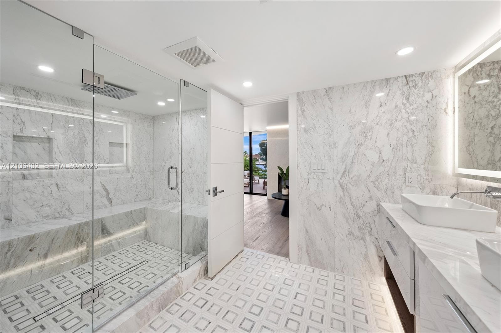Walk-in shower with stone wall cladding, built-in stone bench, and frameless glass enclosure; vanity featuring dual vessel sinks, stone countertops, and back-lit mirrors