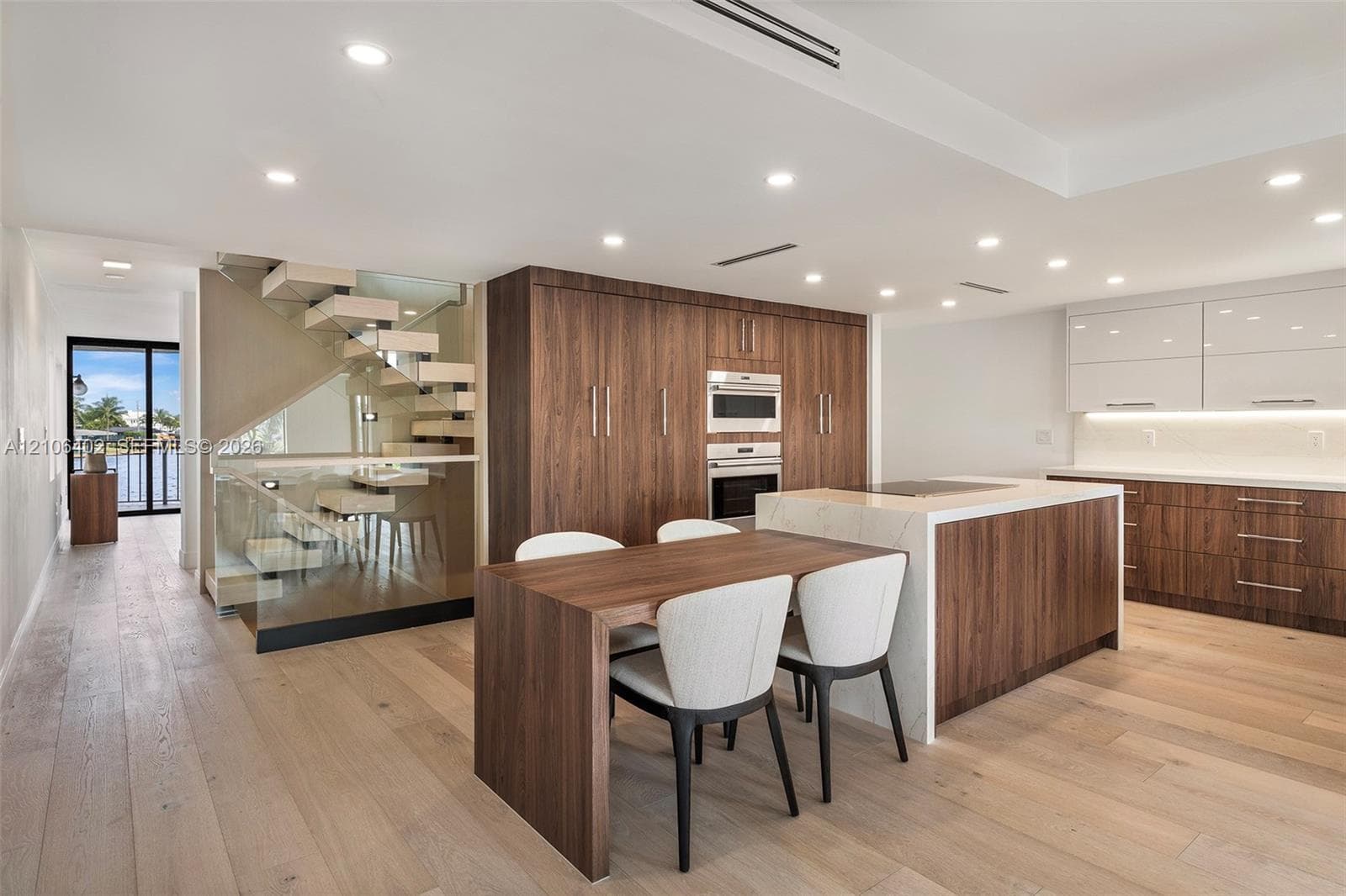 Modern kitchen featuring wood-veneer cabinetry, stone-topped center island with integrated cooktop, recessed lighting, wood-finish flooring, and a floating staircase with glass paneling