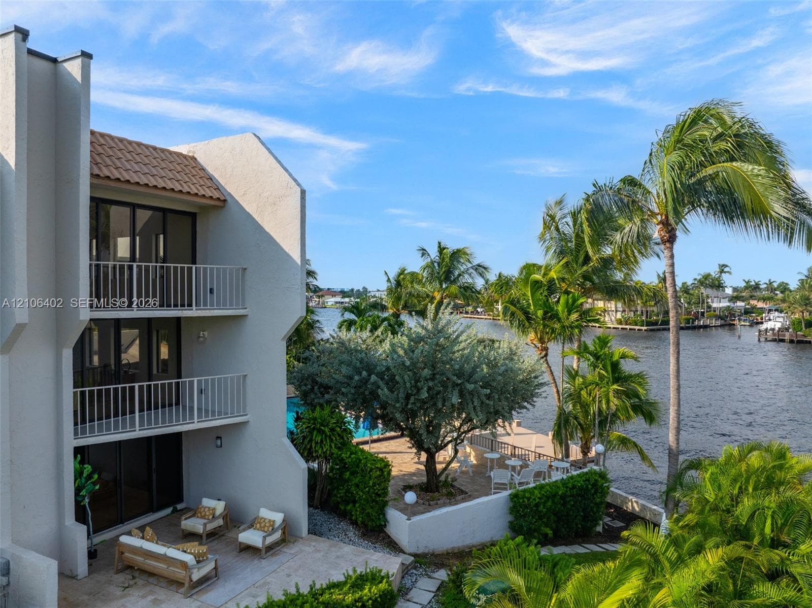 Multi-level exterior featuring light-toned stucco, dual stacked balconies with metal railings, a ground-level stone patio, and direct waterfront access