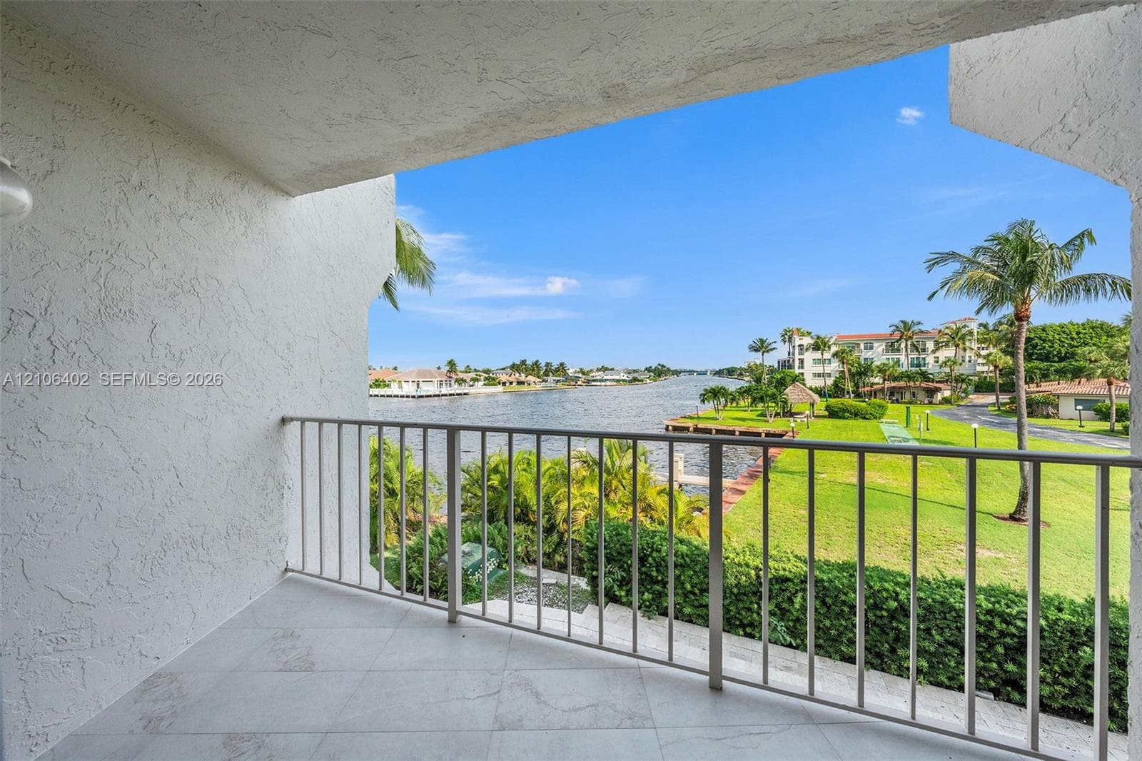Covered balcony featuring neutral large-format tile flooring and white metal railing, overlooking a waterway with manicured landscaping and mature palms