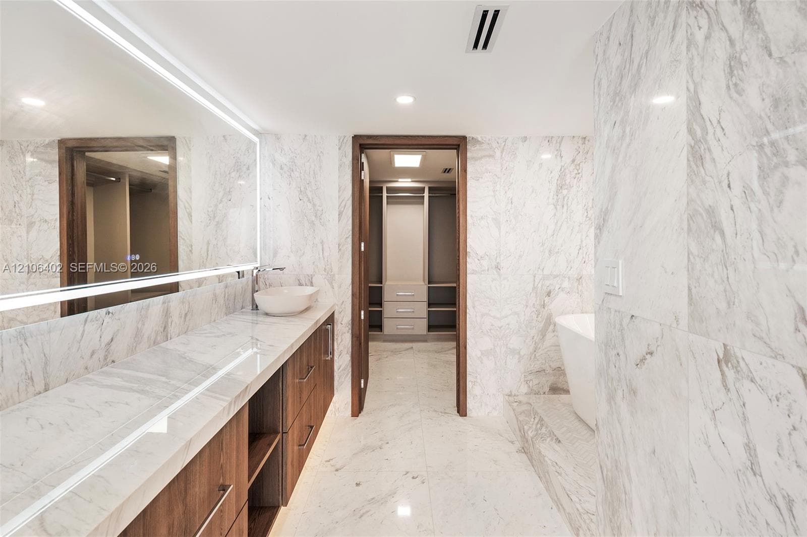 Stone wall and flooring finishes featuring floor-to-ceiling surfaces, wood-finish cabinetry, a vessel sink, an integrated light mirror, and a built-in closet system