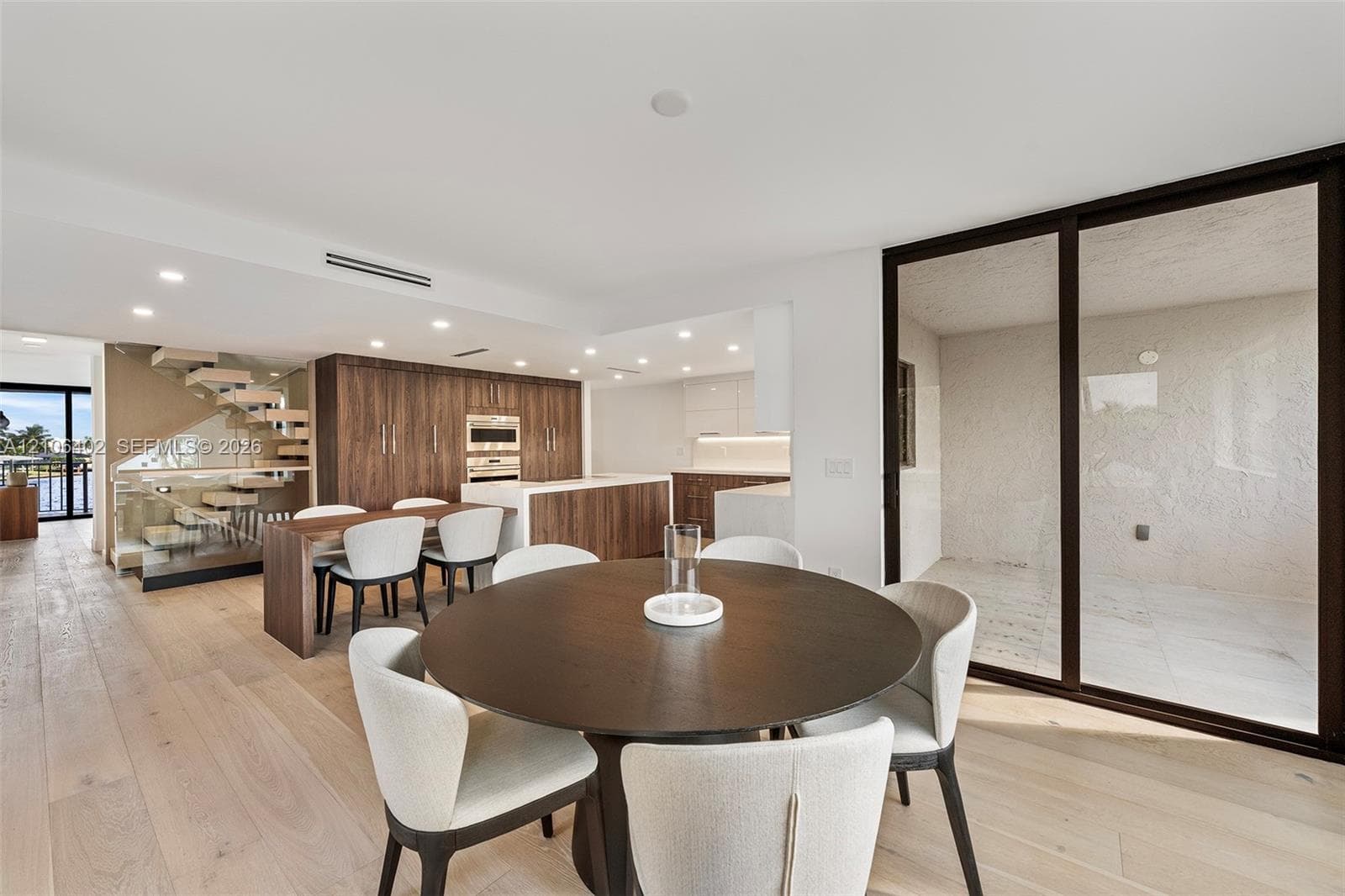 Open-concept interior featuring light wood-finish flooring, floor-to-ceiling dark-trimmed sliding glass doors, built-in wood cabinetry, a center kitchen island with integrated appliances, and a floating staircase with glass railings