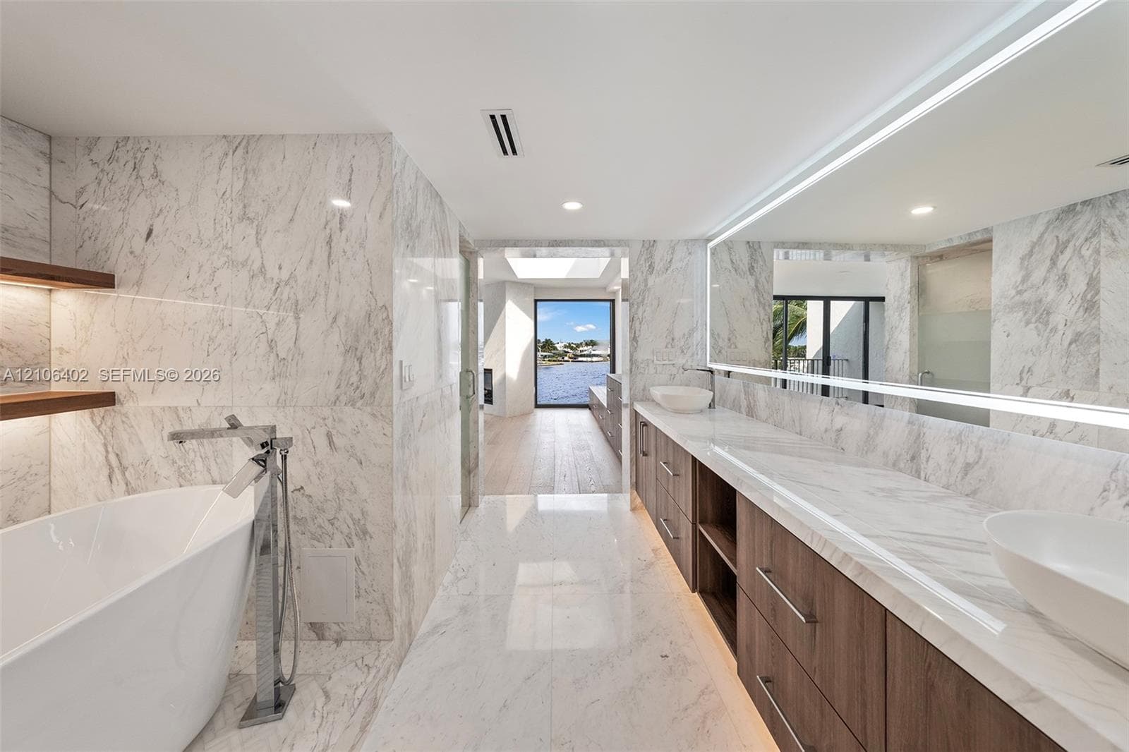 Bathroom featuring stone wall cladding, dual floating vanities with vessel sinks, a freestanding soaking tub, and an expansive integrated mirror with perimeter lighting