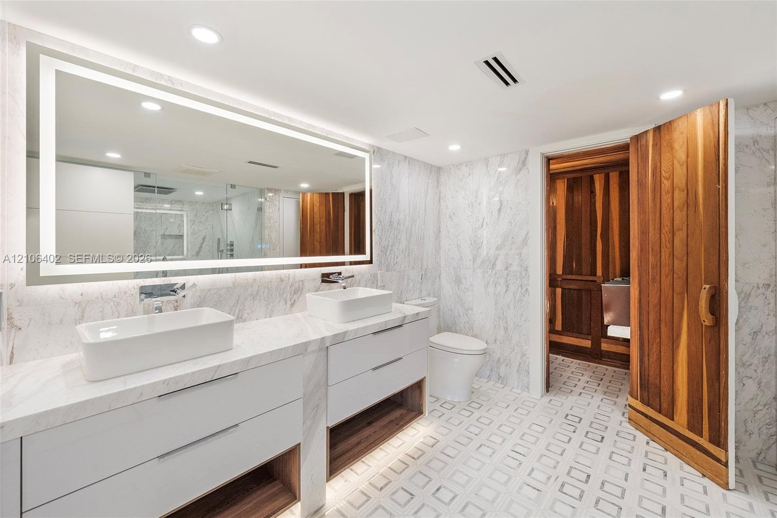 Dual-vanity cabinet with stone countertop and rectangular vessel sinks, large illuminated mirror, stone wall tile, geometric patterned flooring, and wood-paneled sauna entrance