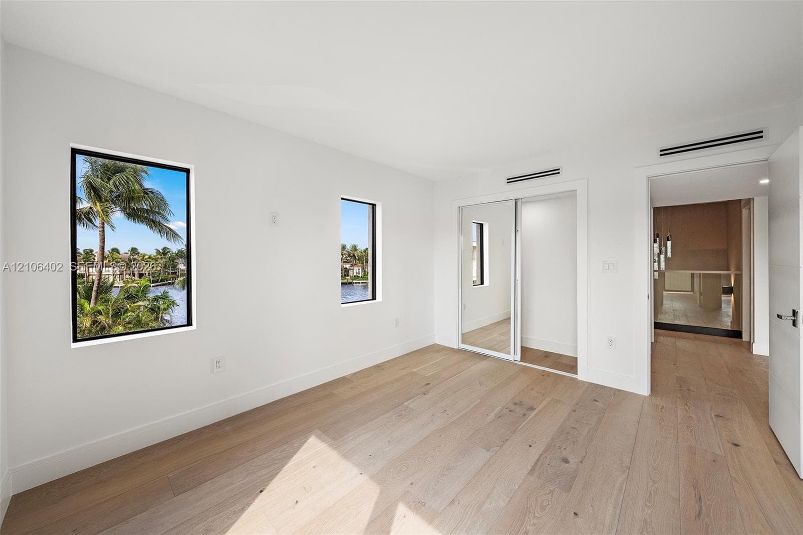 Interior room featuring wide-plank wood-finish flooring, floor-to-ceiling mirror-panel sliding closet doors, and multiple windows with water and palm tree scenery