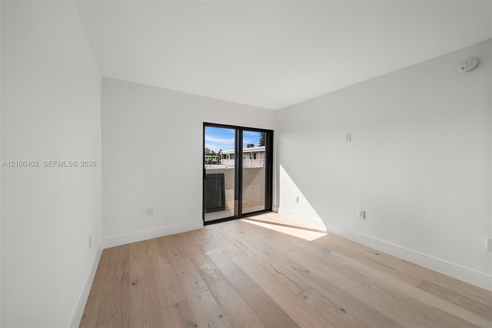 Neutral-toned interior featuring wood-finish flooring, white walls, and black-framed sliding glass doors opening to an exterior balcony