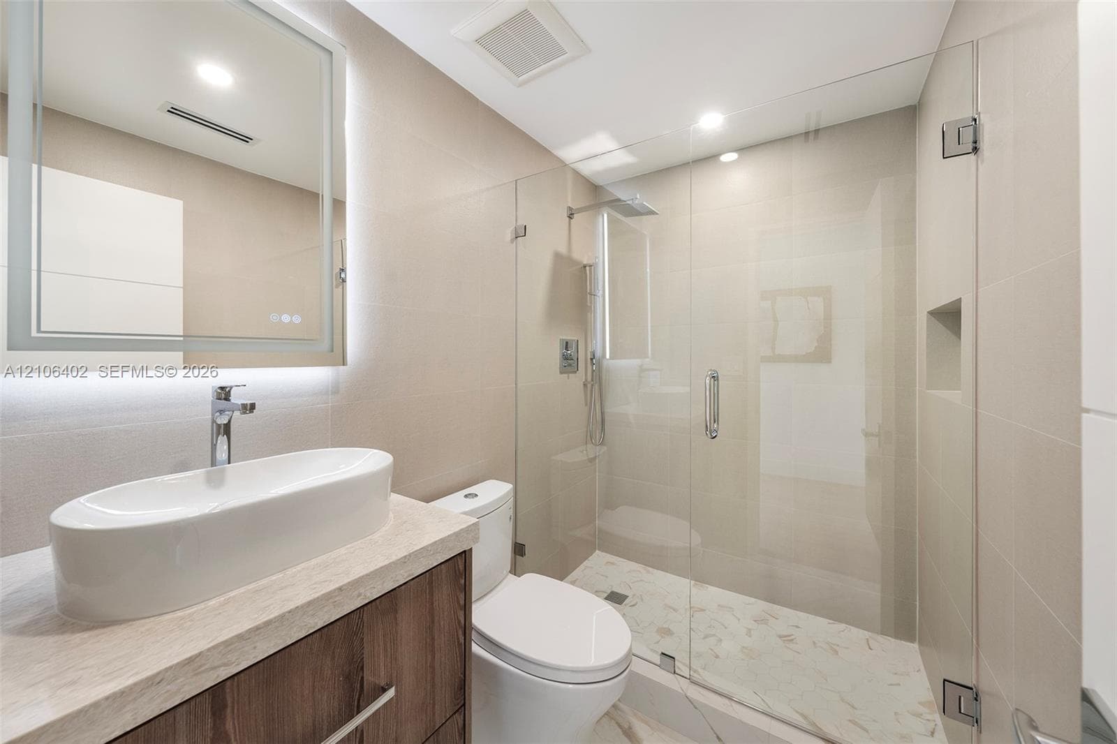 Bathroom featuring a wood-finish vanity with a vessel sink, quartz countertop, illuminated wall-mounted mirror, and a walk-in shower with a glass enclosure and built-in wall niche