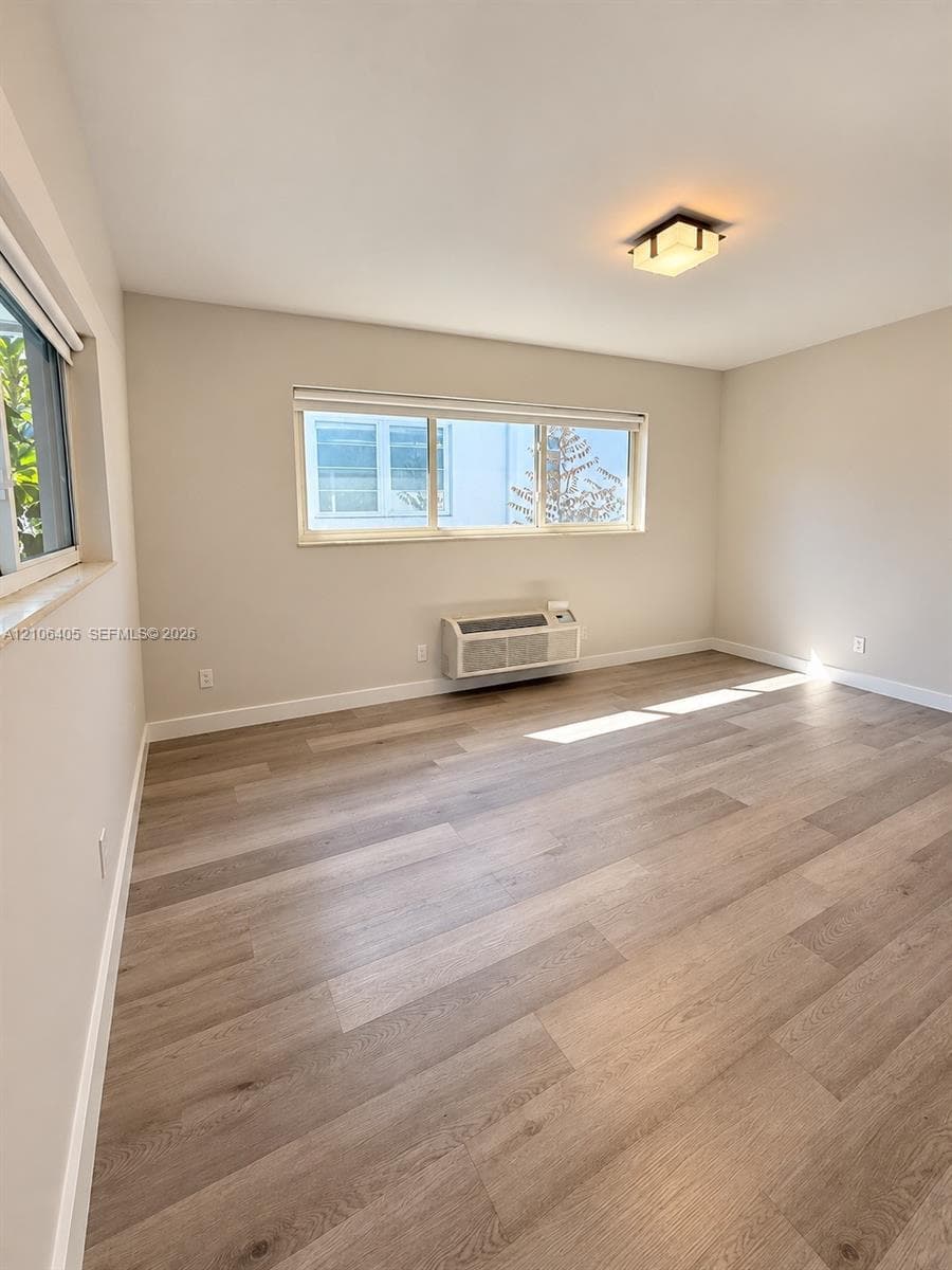 Neutral-toned interior featuring wood-finish flooring, dual horizontal sliding windows, a wall-mounted climate control unit, and a square flush-mount light fixture