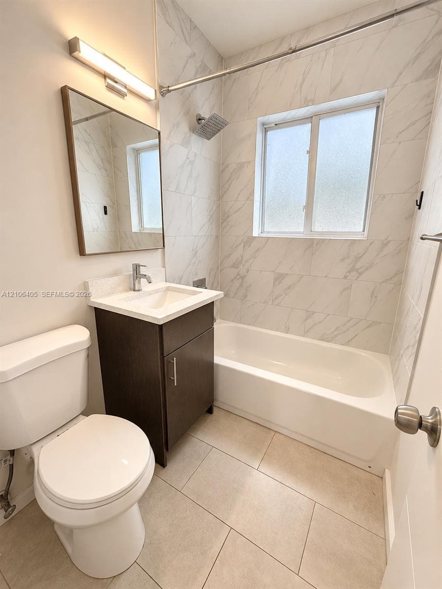 Bathroom featuring a dark wood-finish vanity with a light-toned solid surface countertop, white tile tub surround with veining, large-format floor tile, and rectangular vanity mirror with integrated lighting