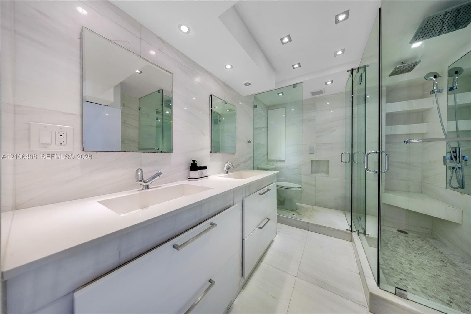 Dual-vanity cabinet with quartz countertops and integrated sinks, glass-enclosed shower featuring a rainfall showerhead, handheld fixture, and built-in bench seating, complemented by recessed lighting and large-format tile flooring