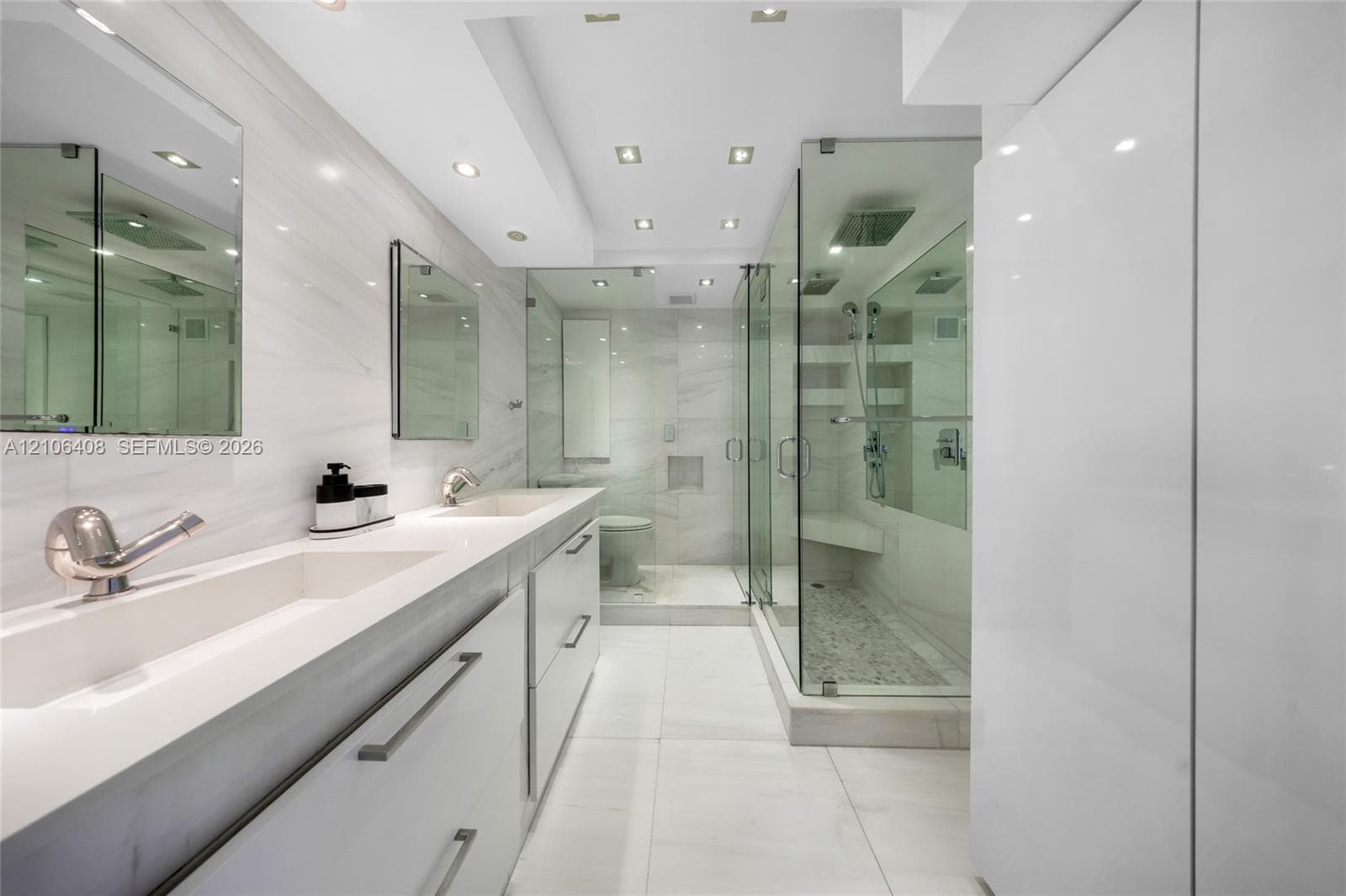 Dual-sink vanity with integrated basins and stone-finish countertops, glass-enclosed shower with built-in bench seating, large-format tile flooring, recessed lighting, and expansive wall-mounted mirrors