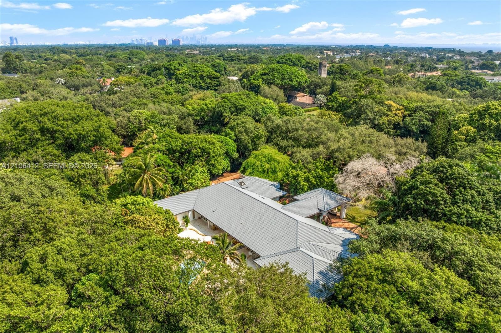 Aerial perspective of a sprawling single-story residence with a metal roof, situated on a heavily wooded lot featuring mature trees, a swimming pool, and a multi-level outdoor patio