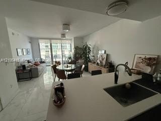 Open-concept interior featuring light-toned tile flooring, solid-surface kitchen countertops, an undermount sink with a gooseneck faucet, and floor-to-ceiling windows