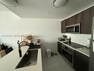 Kitchen featuring dark-toned cabinetry, quartz countertops, an undermount sink with a high-arch faucet, integrated stainless steel appliances, and large-format tile flooring
