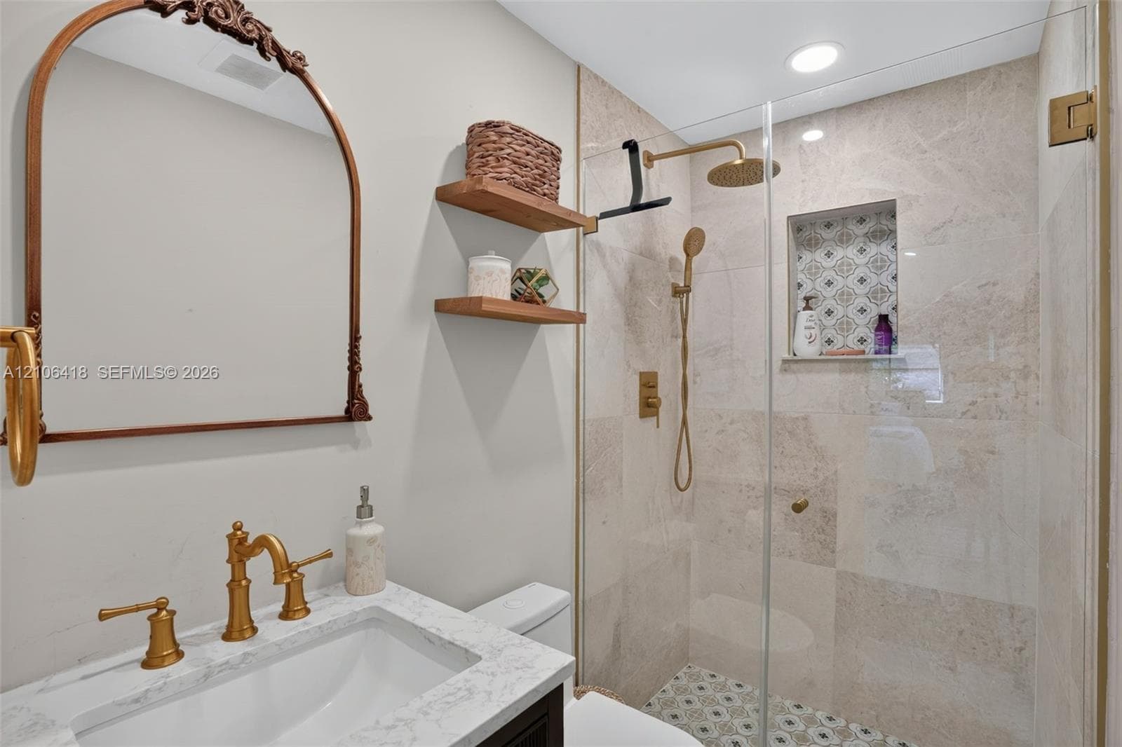 Bathroom featuring an arched metal-frame mirror, stone-composite vanity countertop, wall-mounted wood shelving, glass-enclosed shower with gold-tone hardware, and decorative tiled shower niche