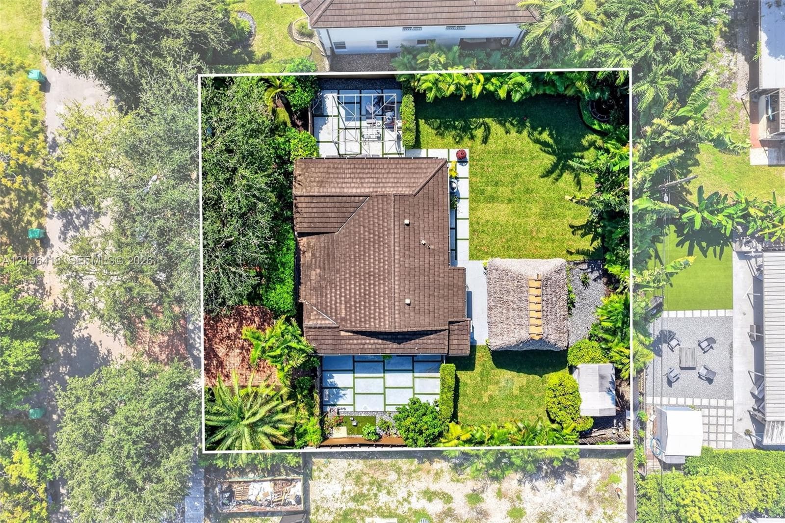 Aerial perspective of a fenced property featuring a tile roof, an enclosed patio area, a detached thatch-roof structure, and extensive landscaped grounds with concrete paver accents