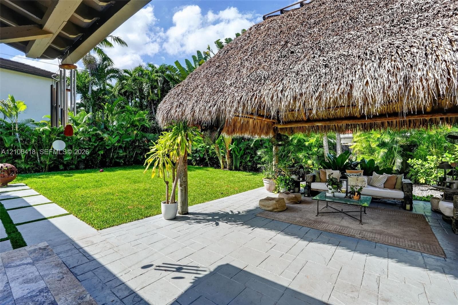 Outdoor living area featuring a natural thatched-roof tiki hut, stone-paver patio, expansive grass lawn, and mature perimeter landscaping