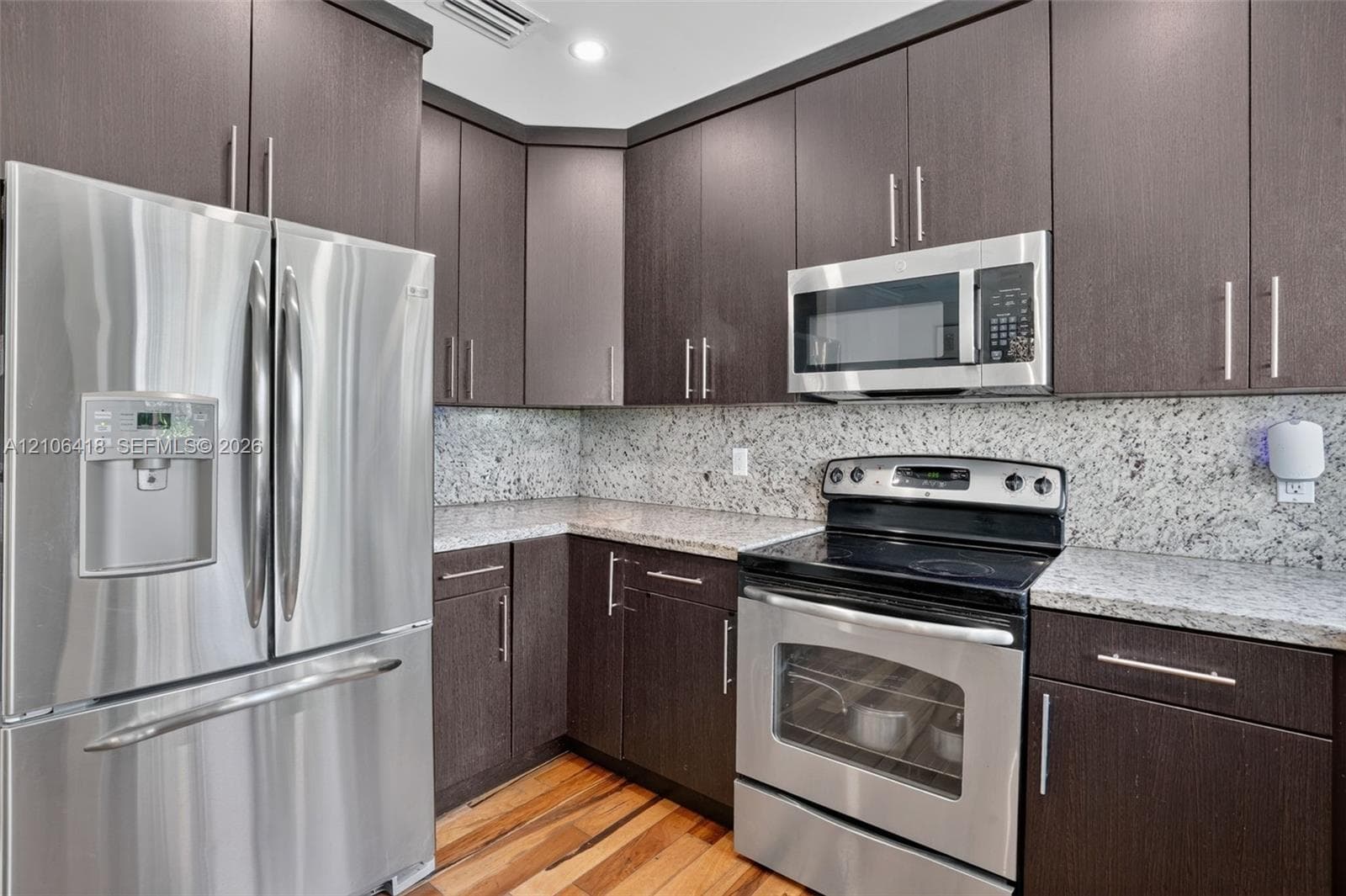 Kitchen featuring dark-toned flat-panel cabinetry, light-colored stone-patterned countertops, stainless steel appliances, and wood-finish flooring