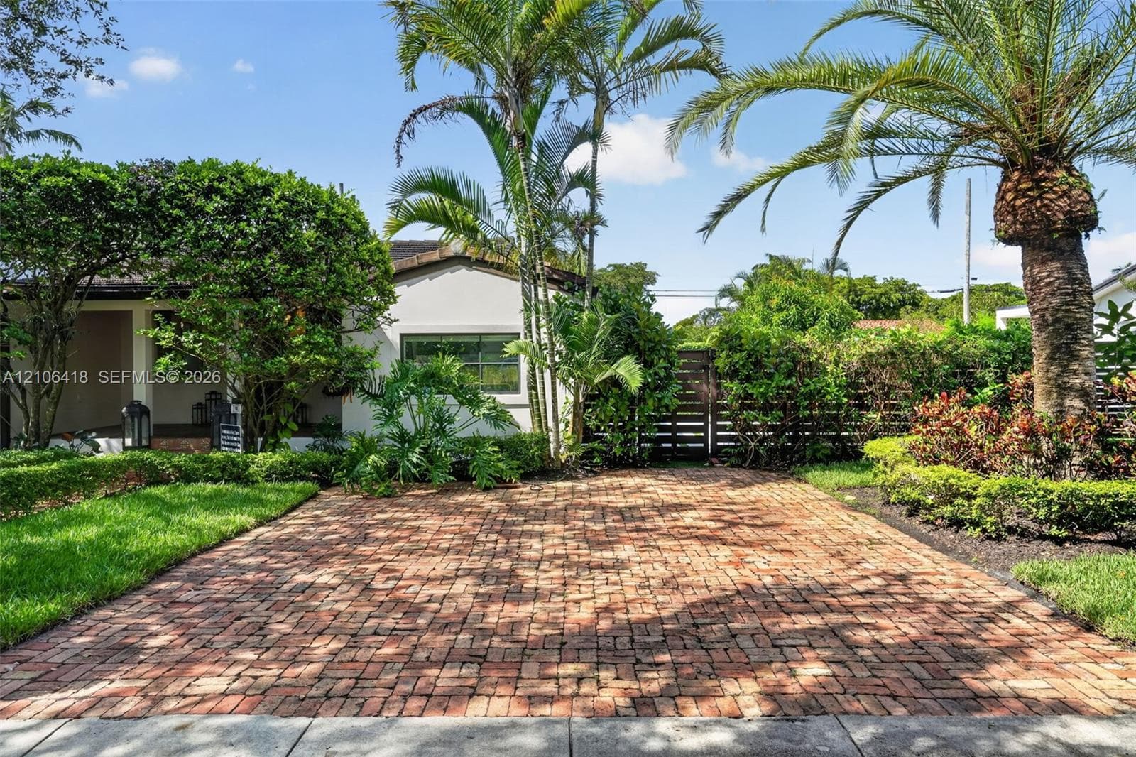 Brick paver driveway, white stucco exterior, mature palm trees, lush manicured landscaping, and dark-toned wooden privacy gate