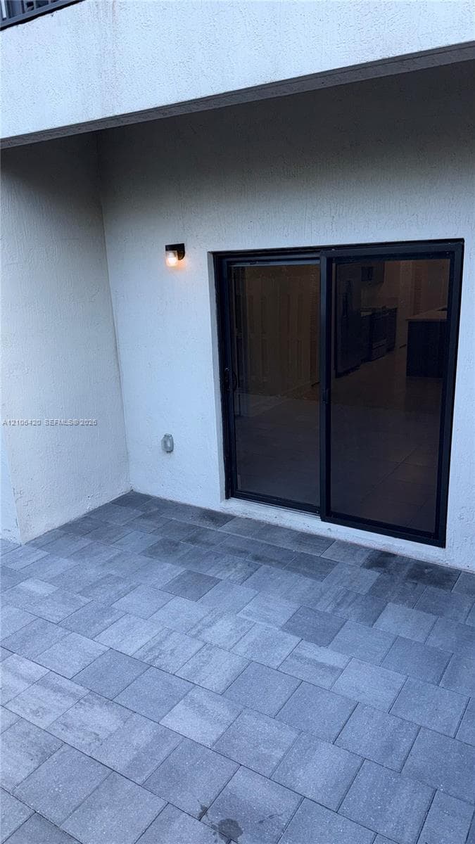 Covered patio featuring gray paver flooring, sliding glass doors with dark frames, and wall-mounted exterior lighting