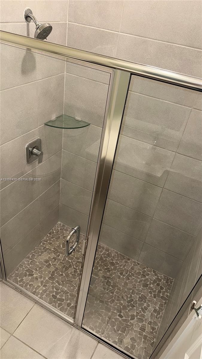 Walk-in shower featuring metallic-finish frame, glass door, pebble-texture stone floor, wall-mounted shower head, and integrated glass corner shelf