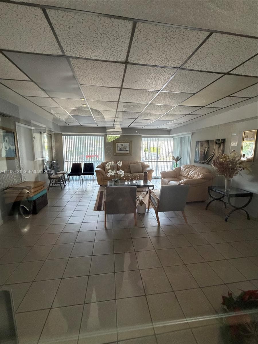 Spacious room featuring neutral ceramic tile flooring, a drop ceiling with integrated panel lighting, and floor-to-ceiling vertical blinds