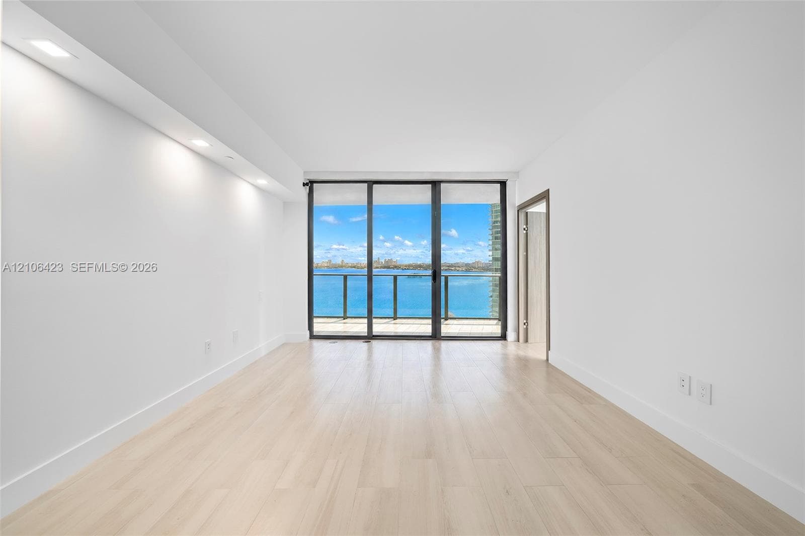 Interior space featuring light-toned wood-finish flooring, recessed ceiling lighting, and floor-to-ceiling glass sliding doors opening to an exterior balcony with water frontage