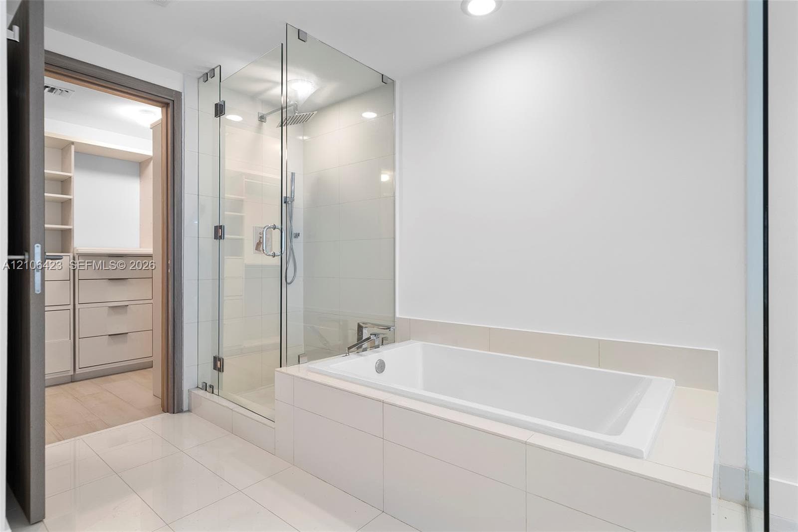 Glass-enclosed walk-in shower with rain showerhead, deep-soaking bathtub with tile surround, built-in custom closet cabinetry, and large-format floor tiling
