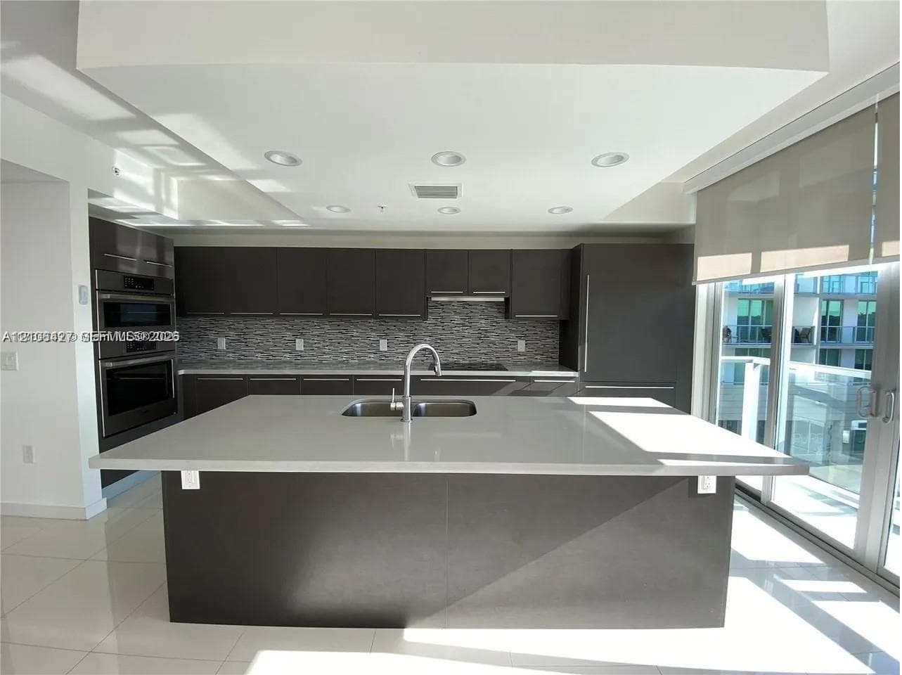 Kitchen featuring dark cabinetry, a large central island with a solid-surface countertop, an undermount sink, built-in stainless steel appliances, and tile flooring
