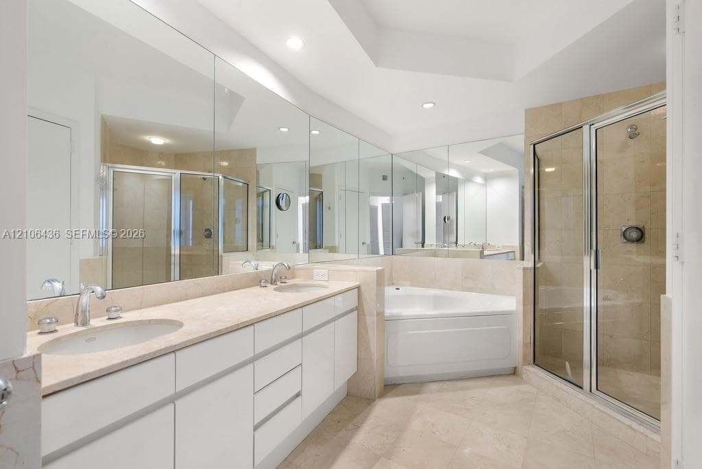 Double vanity with light-toned stone countertop, expansive wall-to-wall mirrors, recessed lighting, corner soaking tub, and enclosed shower with stone-tile surround