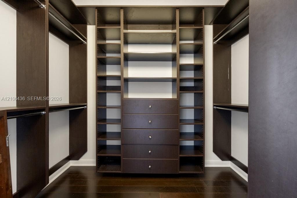 Custom walk-in closet featuring dark wood-finish cabinetry, integrated open shelving, a center bank of five soft-close drawers with metallic pulls, and wood-finish flooring