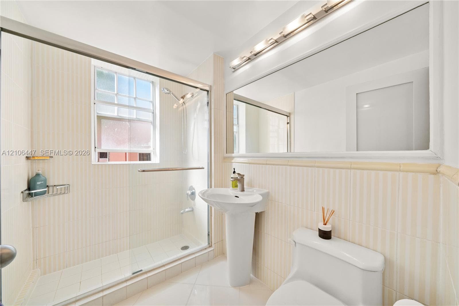 Full-width vanity mirror with integrated multi-bulb lighting, pedestal sink, glass-enclosed walk-in shower with tiled surround, and ceramic tile flooring