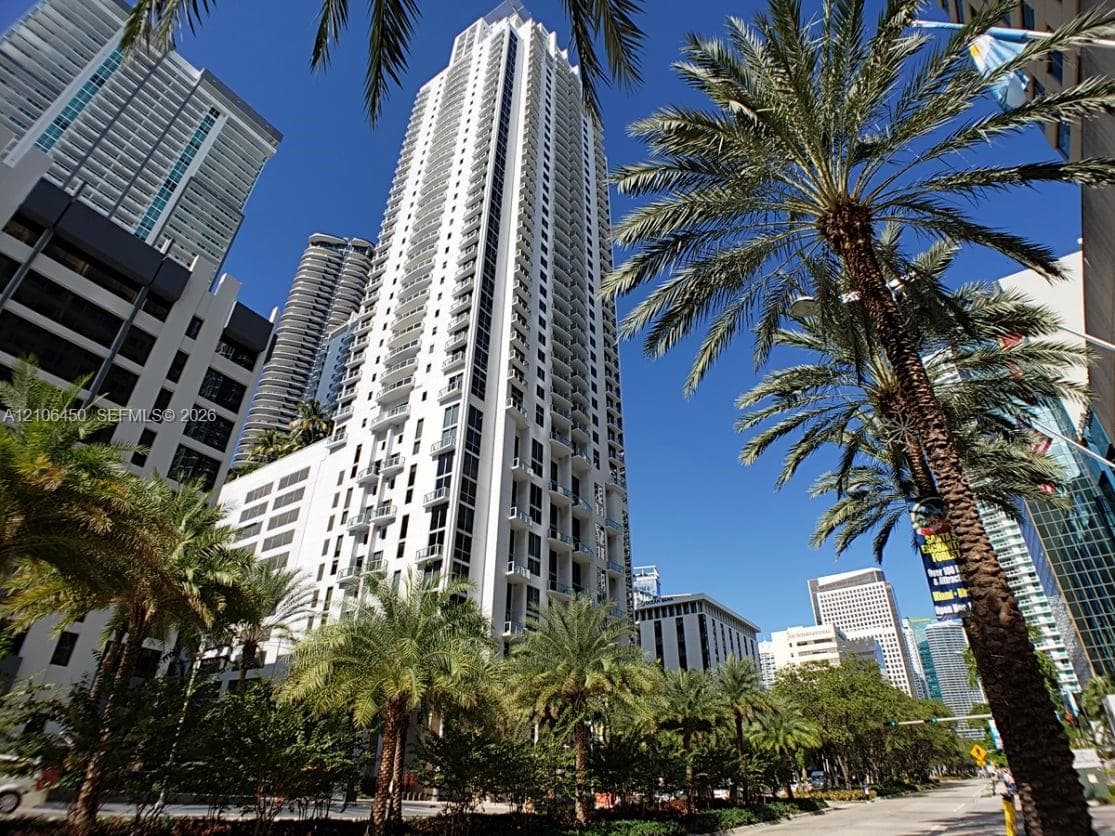 High-rise residential tower featuring a white concrete facade, recessed balconies, floor-to-ceiling windows, and mature palm tree landscaping