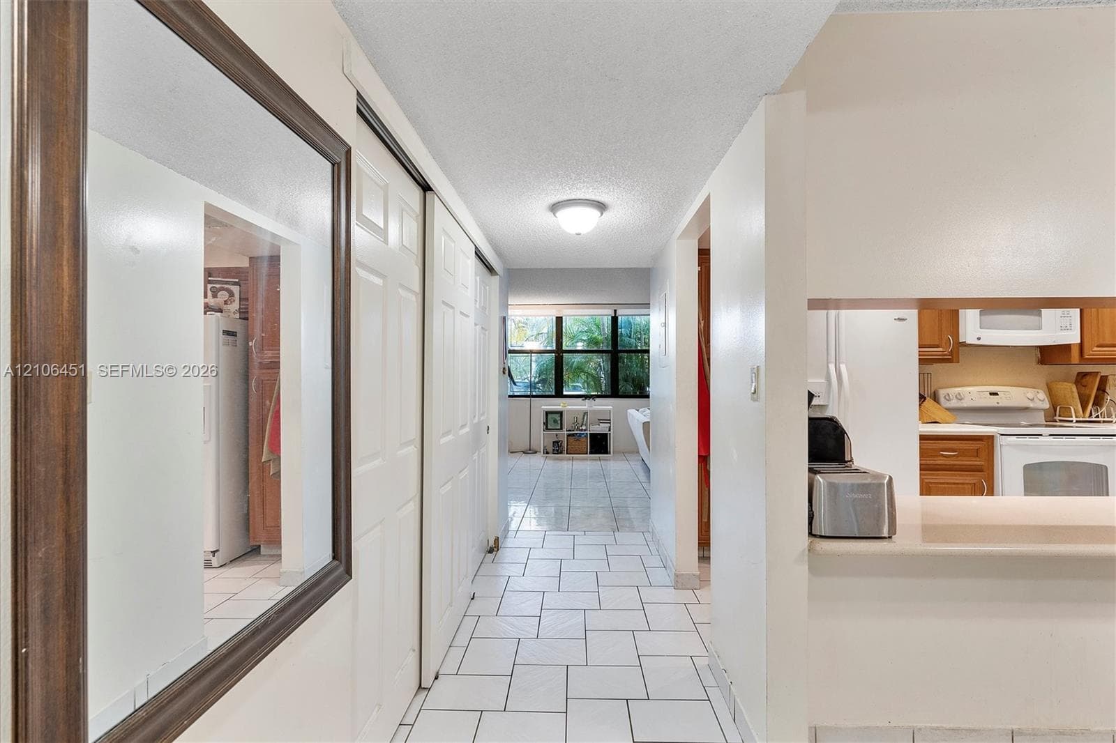 Hallway featuring large-format ceramic tile flooring, double-door closet cabinetry, ceiling-mounted light fixture, and a pass-through kitchen opening with a countertop ledge