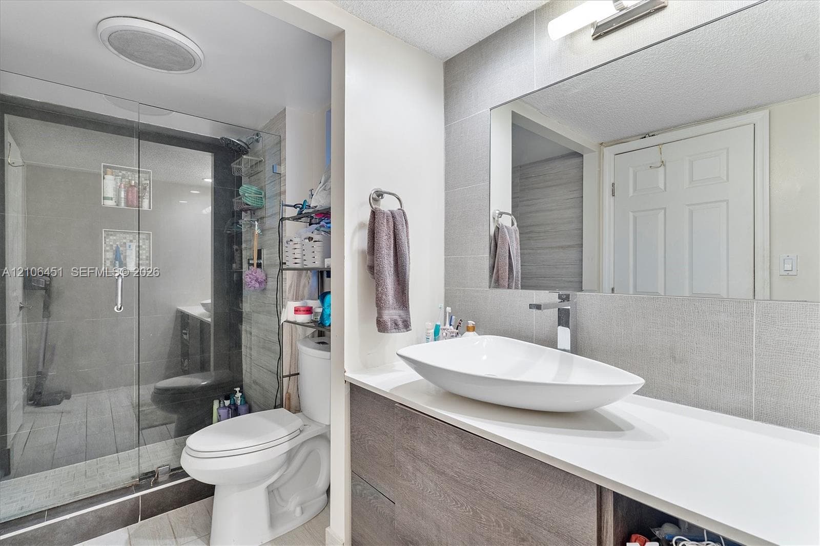Bathroom featuring a wall-mounted vessel sink, wood-finish vanity cabinetry, full-width wall mirror, and a walk-in shower with floor-to-ceiling tile surround and glass enclosure