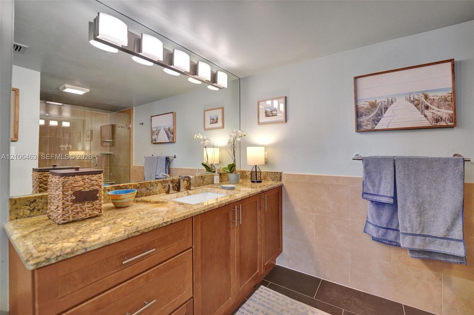 Natural stone countertop featuring an integrated rectangular undermount sink, cherry-tone wood cabinetry with brushed nickel hardware, full-width wall mirror, five-light overhead fixture, and tiled wall wainscoting