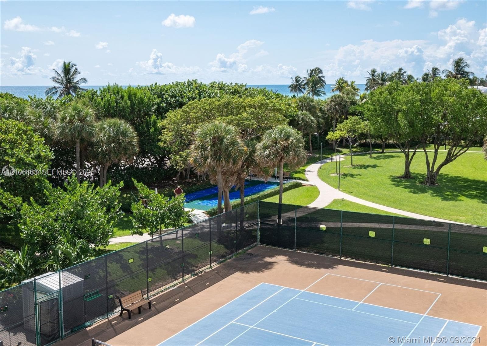 Tennis court with fenced perimeter, paved walking paths, manicured lawns, mature palm trees, and ocean backdrop