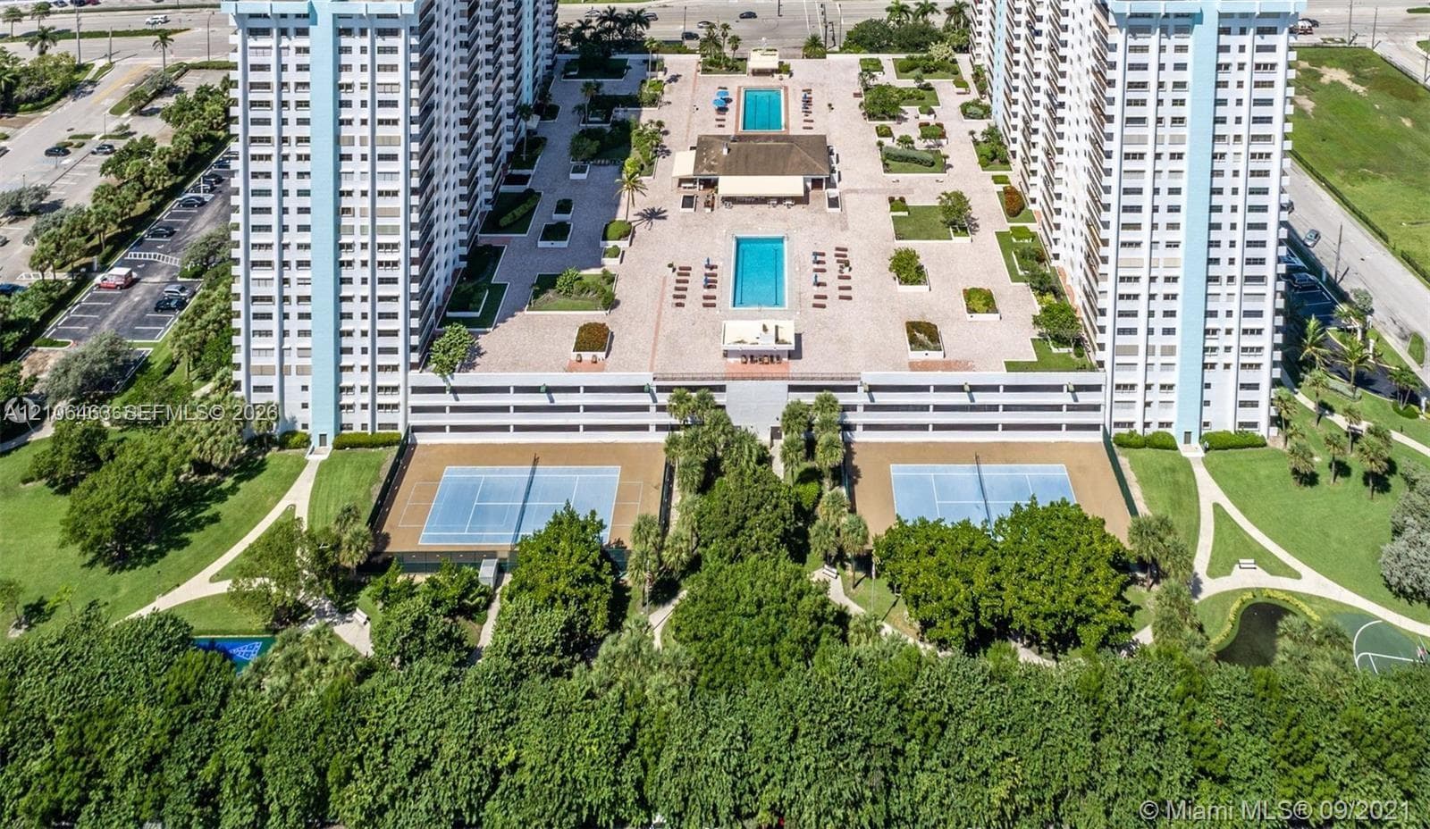 Aerial perspective featuring two outdoor swimming pools, two hard-court tennis courts, extensive paved sun decks, and mature landscaping