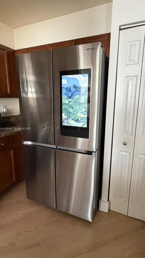 Stainless steel four-door refrigerator with integrated smart display, wood-finish flooring, and dark-toned cabinetry with stone-finish countertops