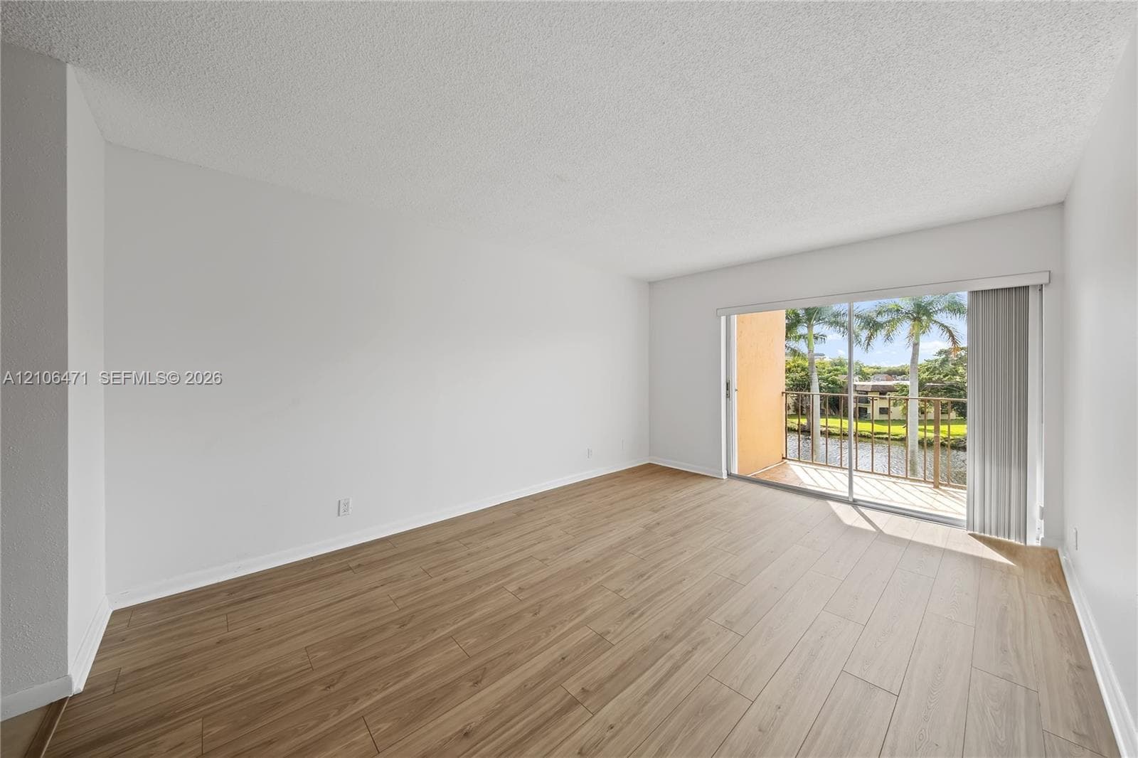 Neutral interior space featuring wood-finish flooring, textured ceiling, and sliding glass doors opening to a balcony with water frontage and palm trees