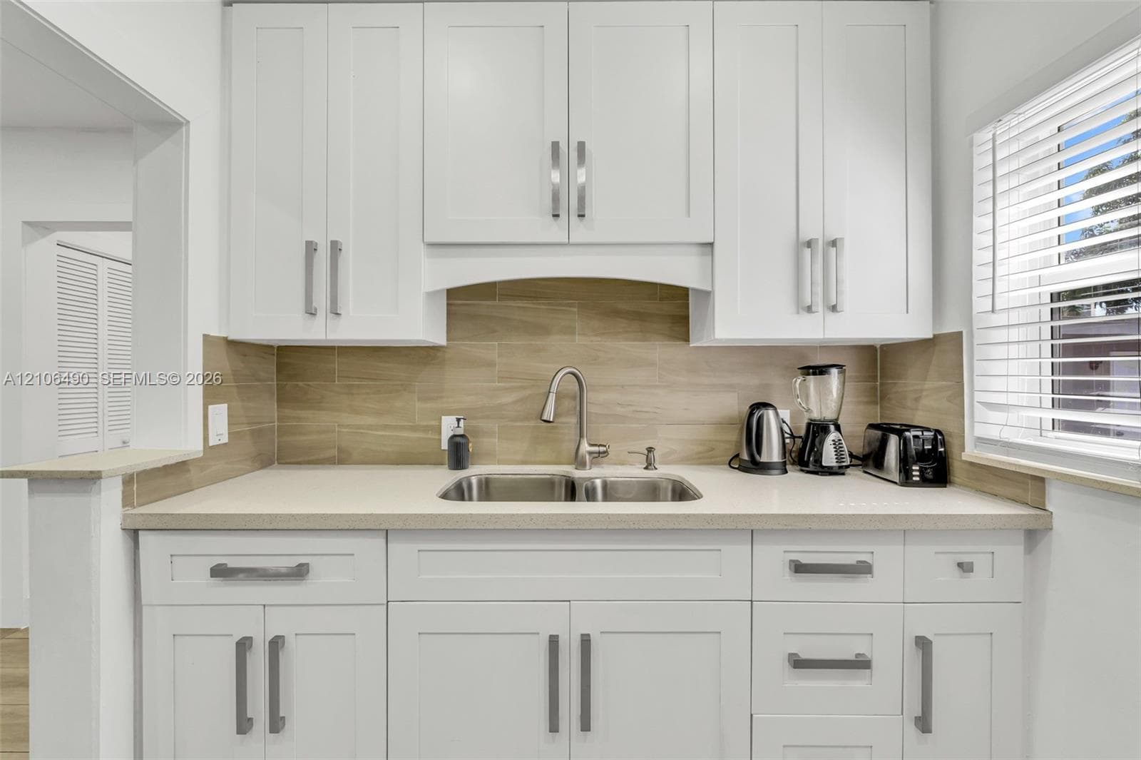 Shaker-style white cabinetry with brushed nickel hardware, neutral solid-surface countertops, horizontal tile backsplash, stainless steel dual-basin undermount sink, and pull-down faucet