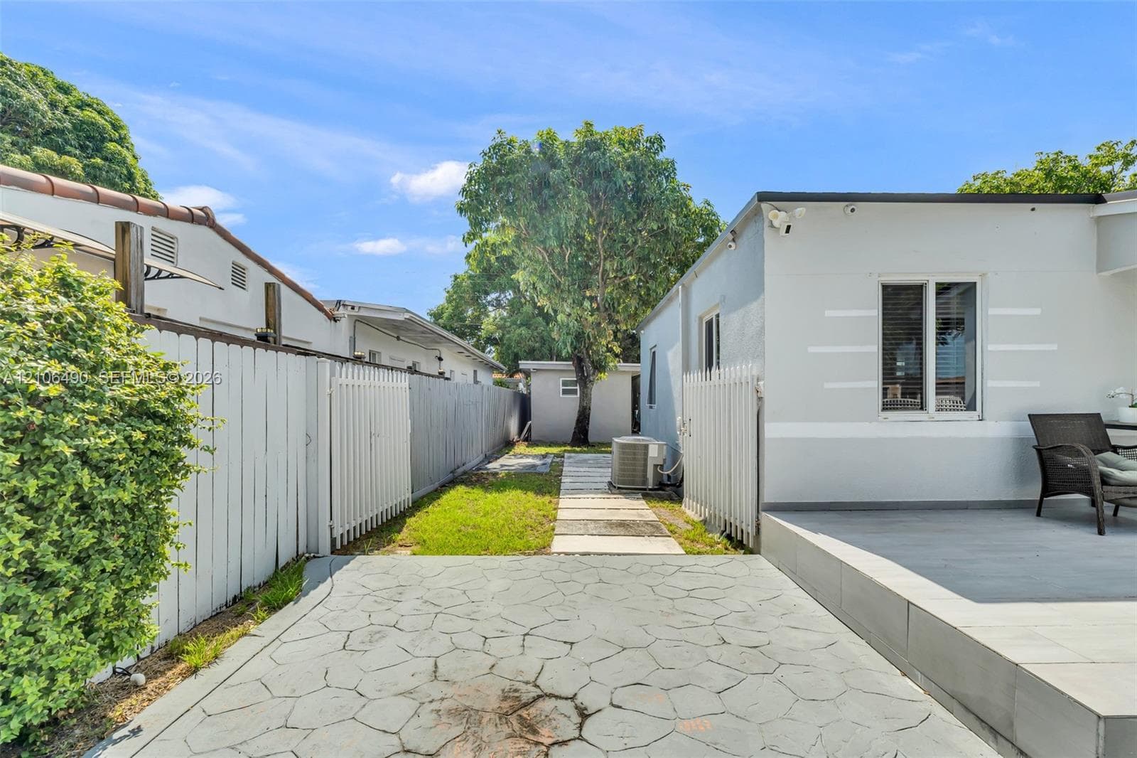 Rear exterior space featuring a stamped concrete patio, paver walkway, white vertical fencing, and mature landscaping