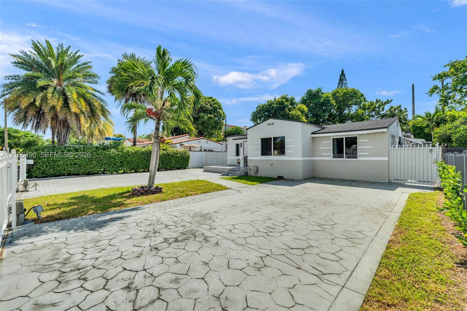 Single-story exterior featuring light-toned stucco siding, a patterned concrete driveway, mature palm trees, and perimeter vinyl fencing