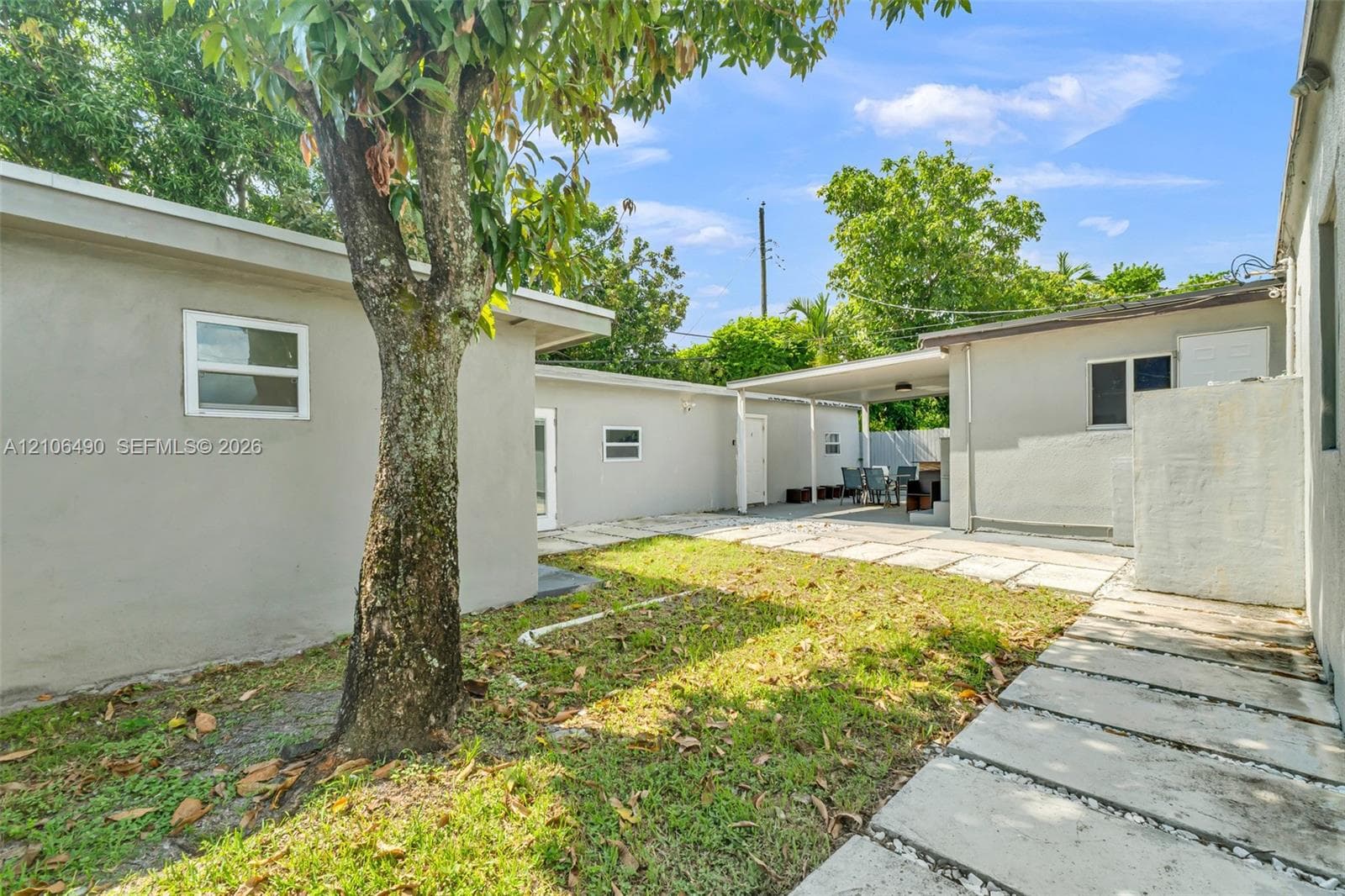 Outdoor space featuring a concrete paver walkway, grassy lawn, mature tree, covered patio, and stucco exterior walls