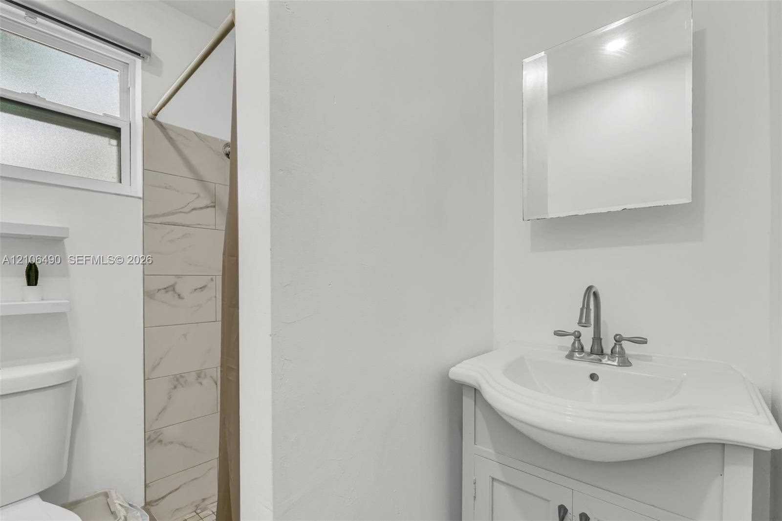 Bathroom featuring a vanity with a white basin and metallic faucet, a wall-mounted rectangular mirror, a shower area with light-toned stone-finish tile surround, and built-in wall shelving