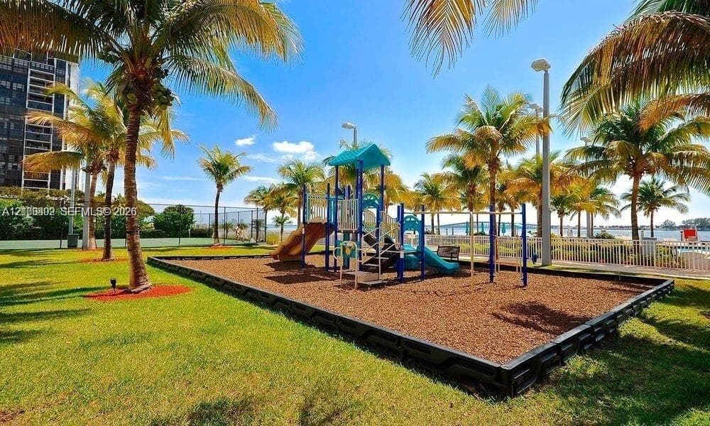 Outdoor recreational area featuring a playground structure with slides, a manicured lawn, established palm trees, and perimeter fencing