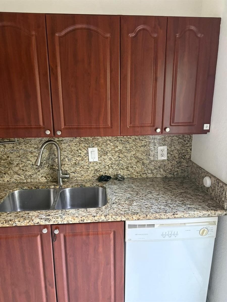 Kitchen featuring arched-panel wood cabinetry, stone-surface countertops, matching stone backsplash, dual-basin stainless steel sink with high-arc faucet, and built-in dishwasher